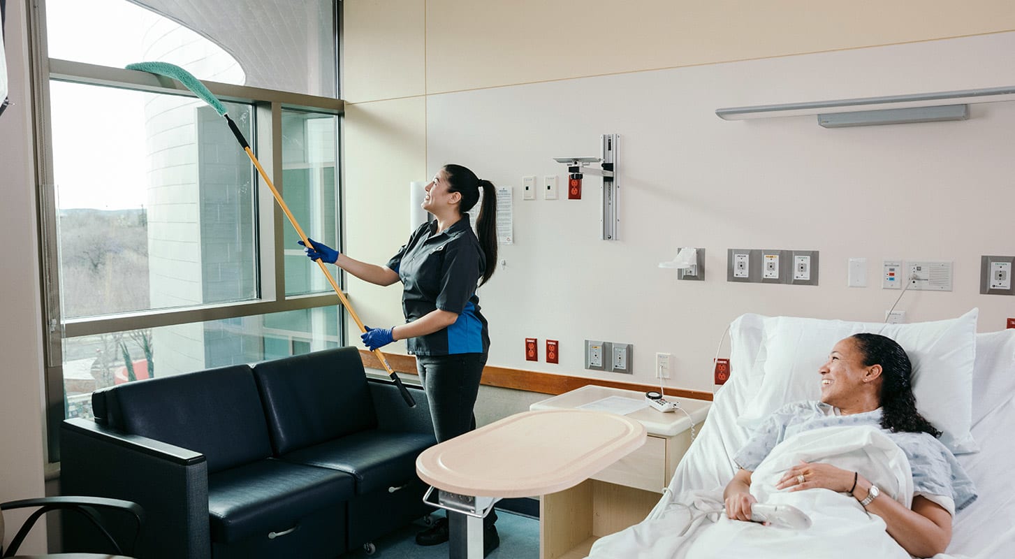 A woman dusts a window in a hospital room while another woman rests in a hospital bed nearby.